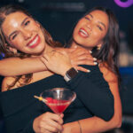 two friends laughing and hugging each other one is holding a drink