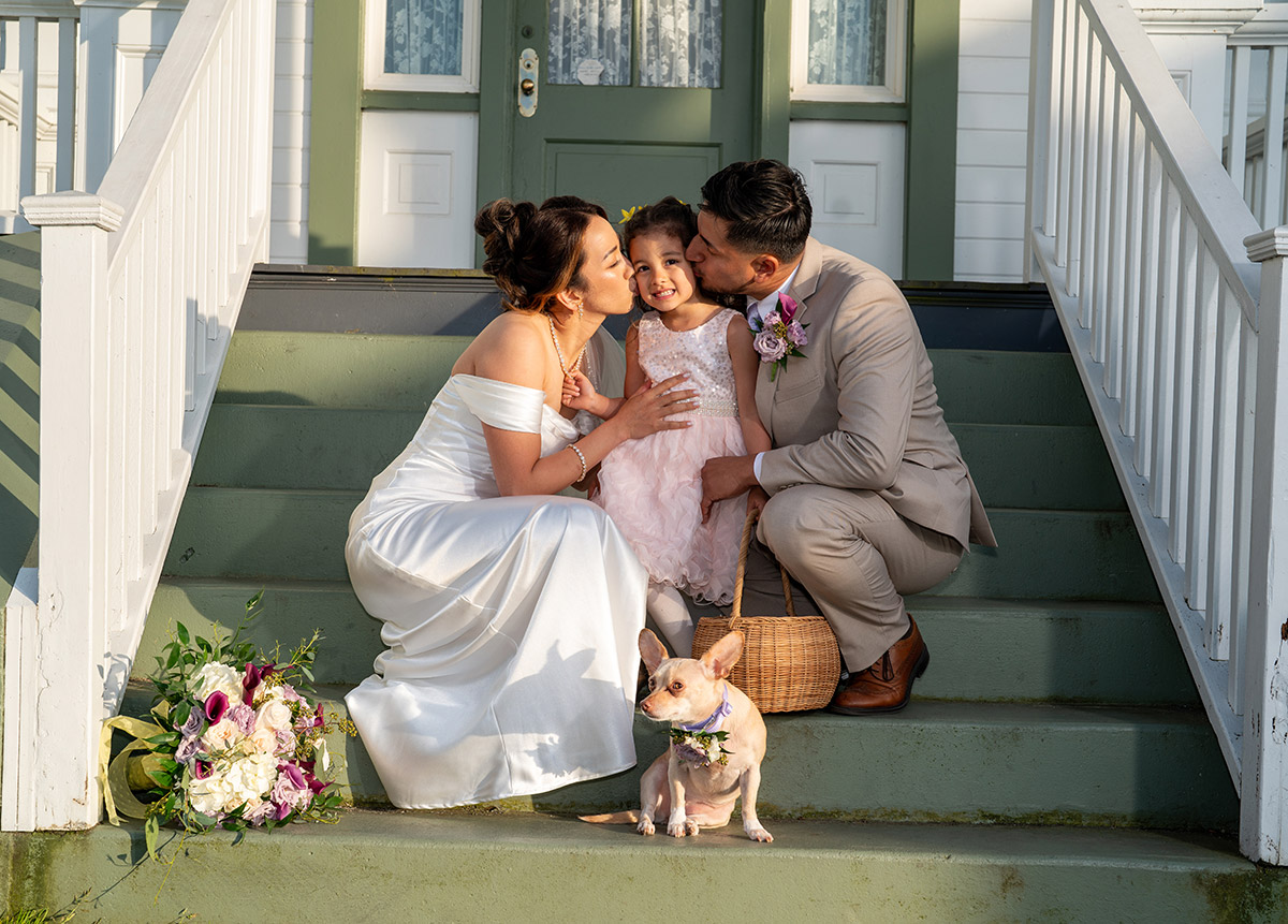 The bride and groom are kissing a kid on the cheek.